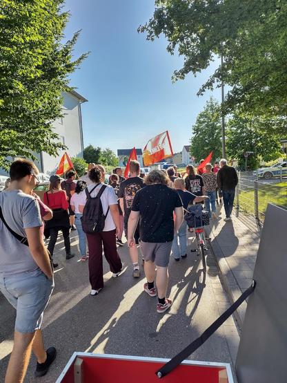 Foto aus einem Demozug heraus, offenbar aus dem Sattel eines roten Lastenfahrrads geschossen. Die Leute tragen rote Linke-Fahnen und gehen unter blauem Himmel auf einer Straße durch ein Wohngebiet mit Bäumen. Rechts hinten ist ein Polizeiwagen zu sehen.