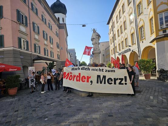 Foto: Auf einem gepflasterten Platz in einer Altstadt-Fußgängerzone hat sich eine Demogruppe um einen Brunnen versammelt, auf dem eine Nepomuk-Statue steht. Der Brunnen ist verdeckt von einem großen Transparent, auf dem steht: „Mach dich nicht zum Mörder, Merz“. Dahinter stehen Menschen, rote Linke-Fahnen wehen. Der Himmel ist blau.