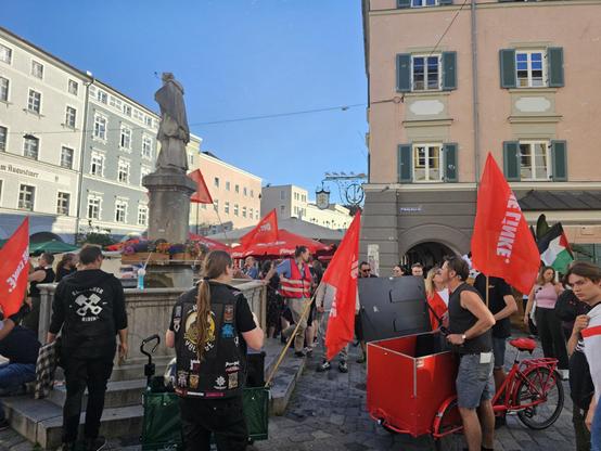 Foto: Ein Demozug macht in einer historischen Altstadt an einem Platz mit Brunnen und Brunnenstatue Station. Es wehen rote Linke-Fahnen, jemand schiebt ein rotes Lastenfahrrad. Links zieht sich eine Häuserreihe in den Hintergrund. Rechts ist ein Haus mit Arkaden, an dem ein Wirtshausschild hängt.