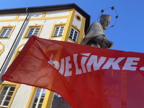 Foto: Blick von unten auf eine steinerne Nepomuk-Statue vor blauem Himmel, gut erkennbar am Sternenkranz um den Kopf. Sie blickt nach unten und scheint mit der Hand nach einer roten Fahne zu greifen, die groß im Vordergrund weht. Darauf steht: Die LINKE. Im Hintergrund ragt schräg die gelb-weiße Fassade eines mehrstöckigen historischen Gebäudes herein.